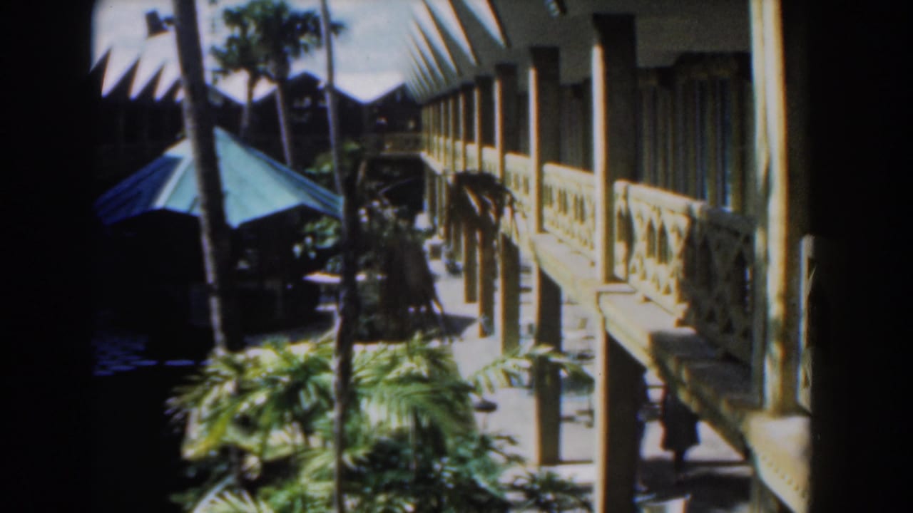 Exterior walkway with yellow railings, palm trees, garden shade, and a small figure at right.