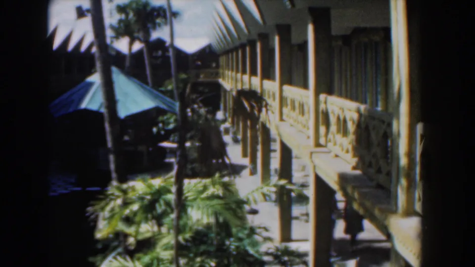 1961 Nassau frame of a bright resort-like exterior with railings, palms, a green umbrella, and a person walking in shadow.