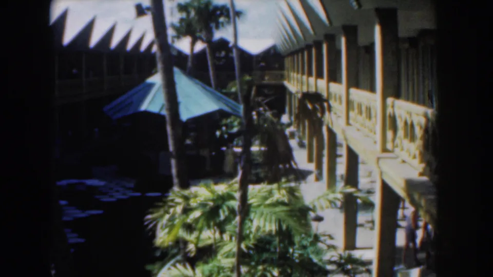 1961 Nassau 8mm frame showing a palm-lined courtyard, blue-green umbrella, exterior railings, and visitors partly visible near the lower edge.