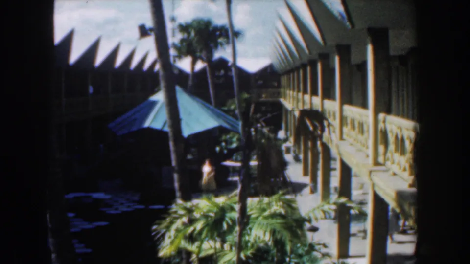 1961 Nassau travel frame with a long exterior walkway, tropical plantings, green umbrella, and a person passing through shade.