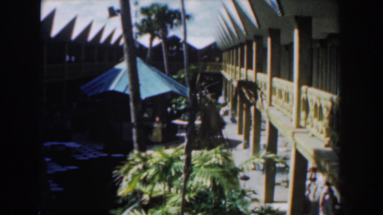 Courtyard with green umbrella, palms, white building, and a person walking in shade.