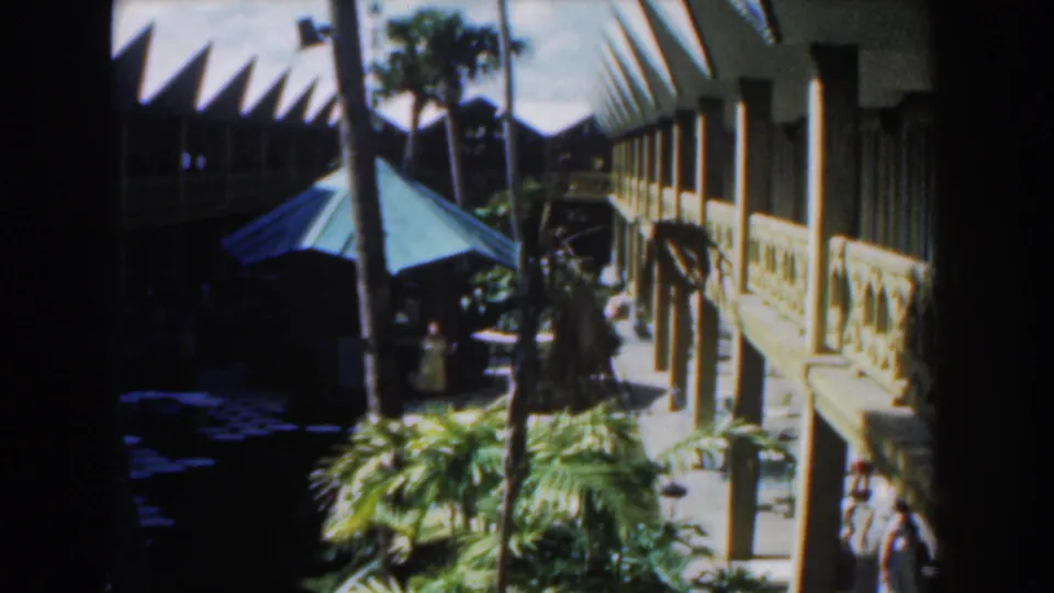 1961 Nassau resort courtyard with a green umbrella, palms, white building, and a distant person in a light dress.