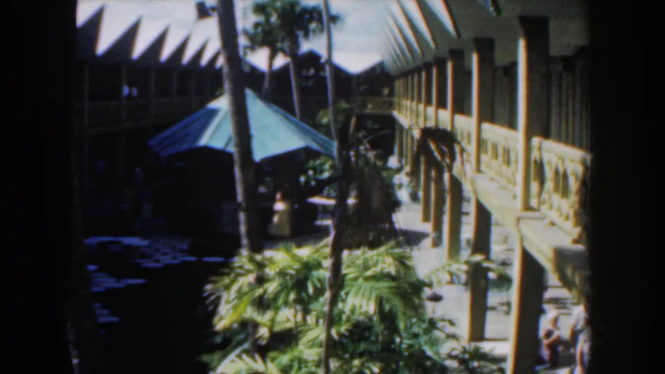 1961 Nassau courtyard frame with a green patio umbrella, white balcony structure, palms, and deep shaded water.