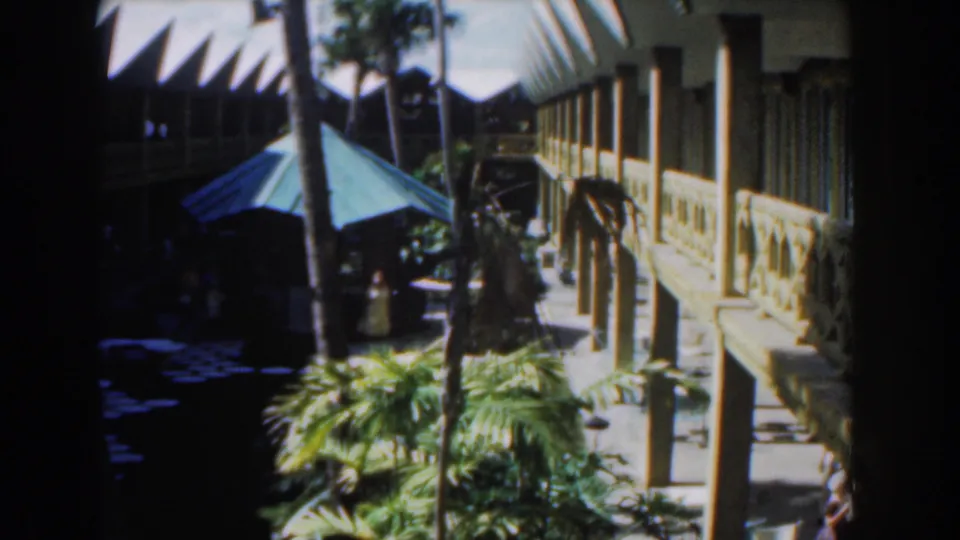 1961 Nassau view of a white resort-style building, green umbrella, palms, and a distant pedestrian in the courtyard.