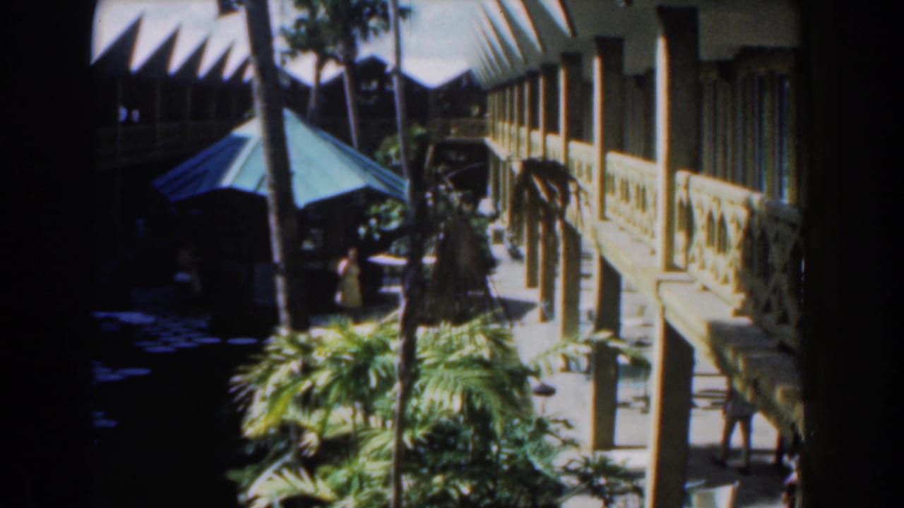 Sunlit courtyard with palms, a blue canopy, and a two-storey building with railings.