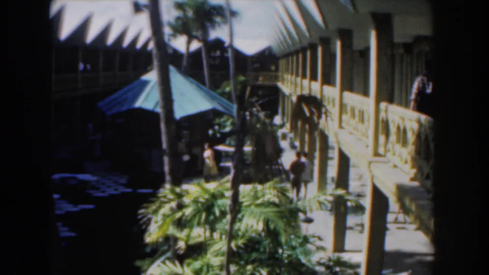 1961 Nassau home-movie still showing visitors in a sunlit courtyard framed by palms, a blue canopy, and a two-storey building.