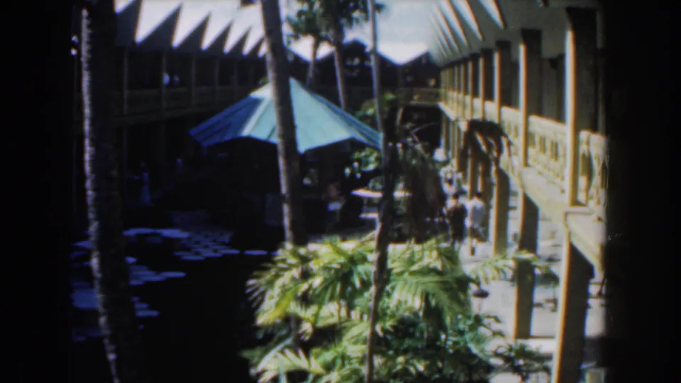 1961 Nassau frame of a courtyard scene with people near the walkway, a blue canopy, palms, and a two-storey building in bright sun.