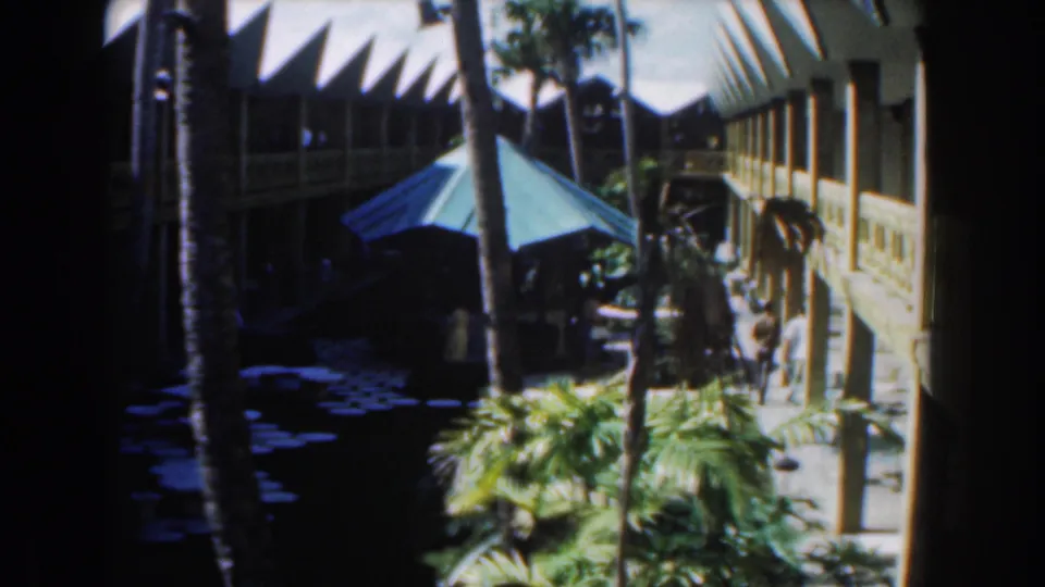 1961 Nassau home-movie view of a tropical courtyard with palms, blue umbrella, shaded paths, and a two-story building.