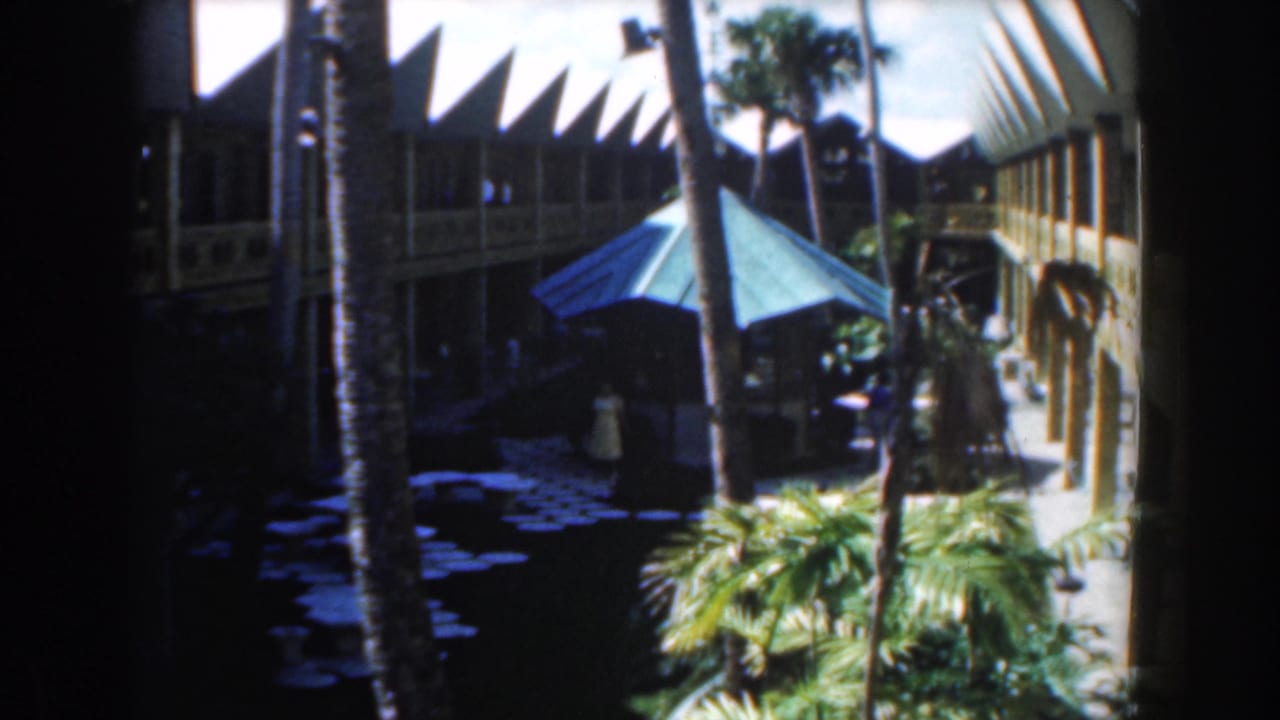 Courtyard view with palms, blue umbrella, foliage, and guests on a walkway.