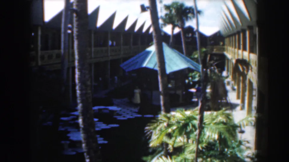1961 Nassau courtyard scene showing palm trees, dense planting, a blue umbrella, and distant guests near resort walkways.