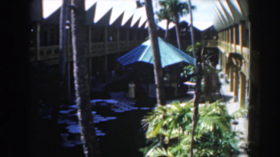 Nassau 1961 travel-film frame with a blue umbrella centered among palms, foliage, shaded paths, and a two-story building.
