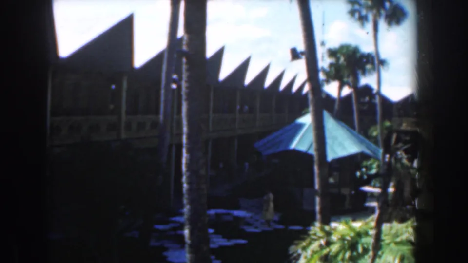 1961 Nassau home-movie frame looking across a tropical courtyard toward angular rooflines, palms, a blue umbrella, and a guest below.