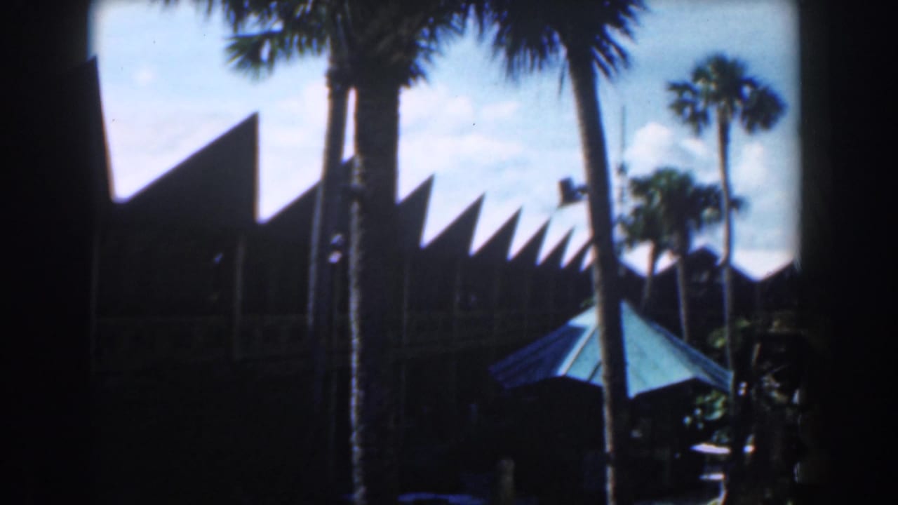 1961 Nassau resort exterior with palm trunks, sharp roof peaks, deep shade, and a turquoise umbrella in the foreground.