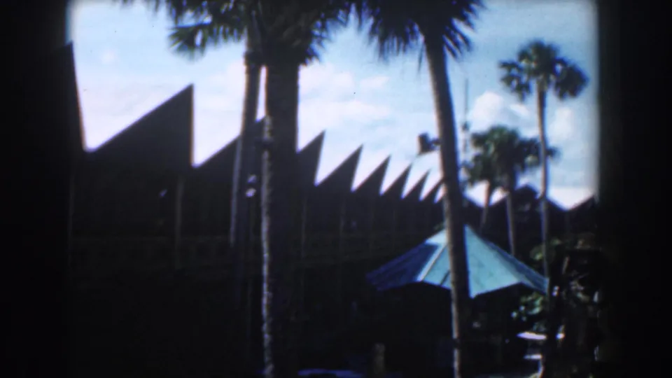 1961 Nassau resort exterior with palm trunks, sharp roof peaks, deep shade, and a turquoise umbrella in the foreground.