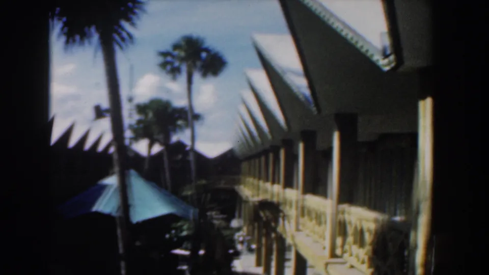 Nassau hotel exterior with palm trees, a shaded walkway, repeated roof points, parked vehicles, and a turquoise umbrella.