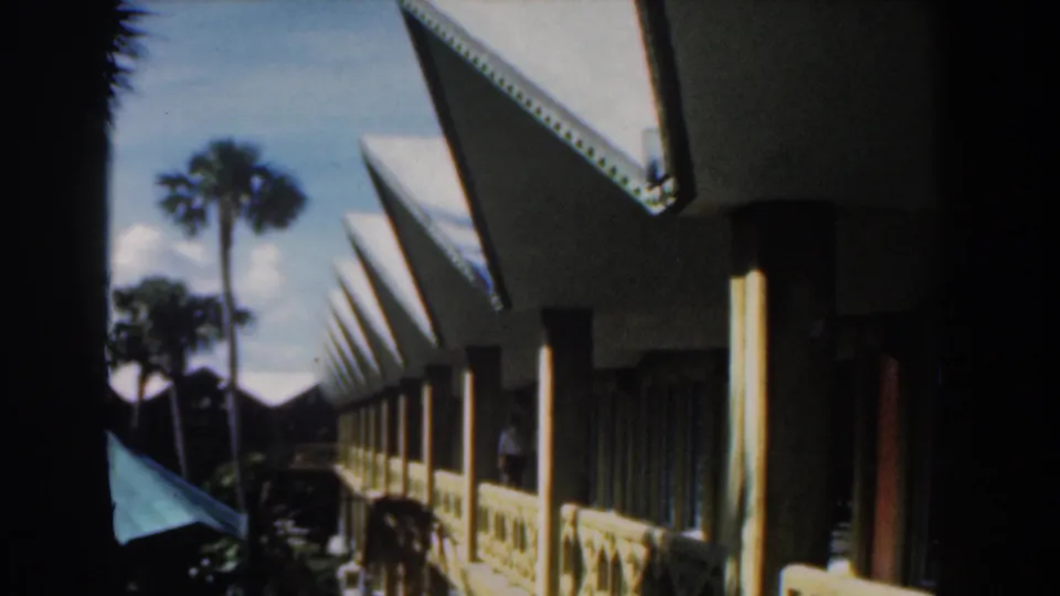1961 Nassau resort walkway with angular roof peaks, palms, a turquoise umbrella, railings, and a distant person near the building.