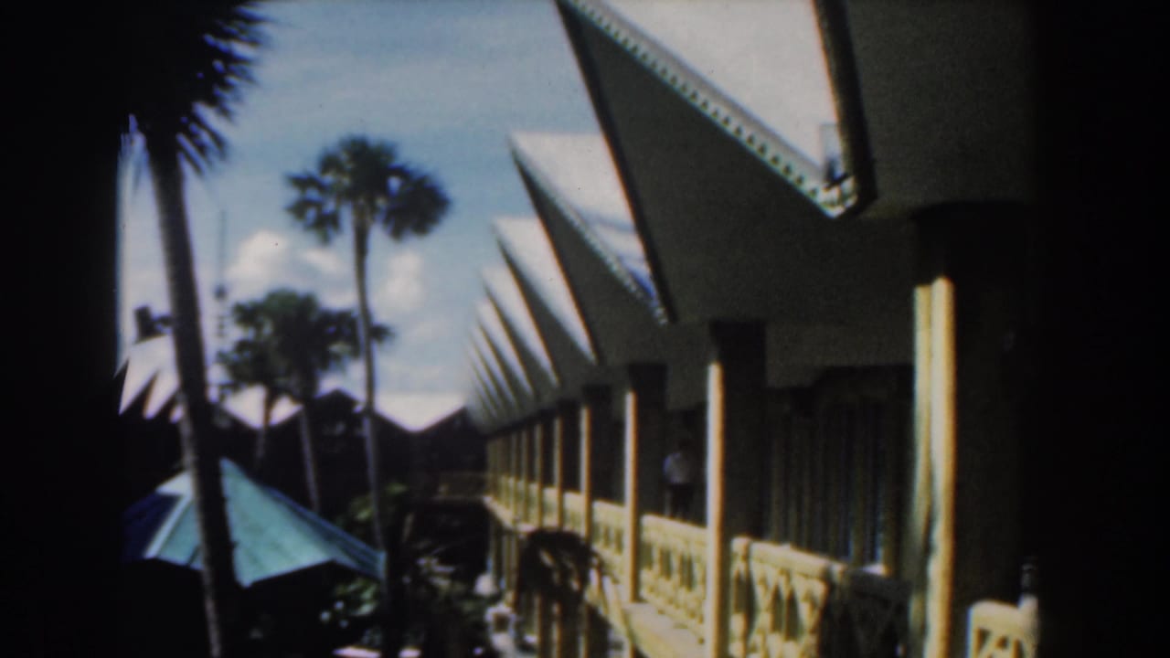 1961 Nassau home-movie view of a white building with pointed roof edges, palms, shaded posts, and a green umbrella.
