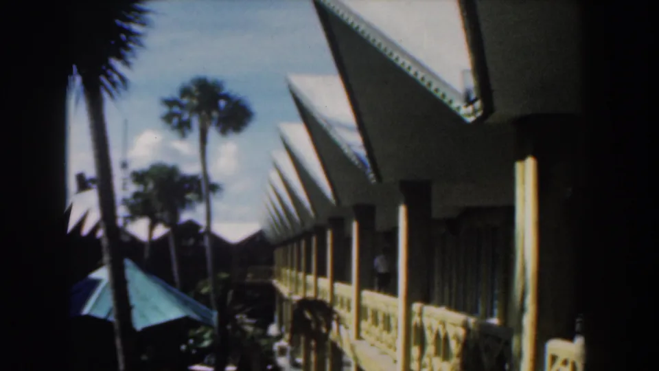 1961 Nassau home-movie view of a white building with pointed roof edges, palms, shaded posts, and a green umbrella.