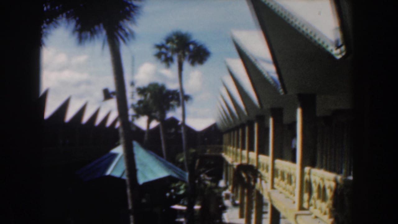 Palm trunks and a green umbrella stand beside a white building with repeating pointed roof sections.