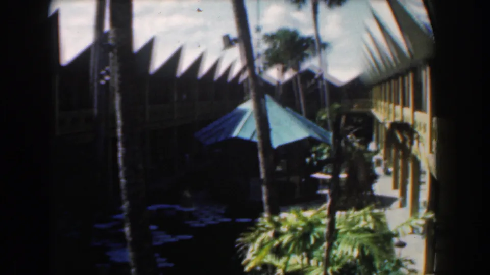 Shaded 1961 Nassau home-movie frame showing palms, courtyard greenery, a green umbrella, and the white building beyond.