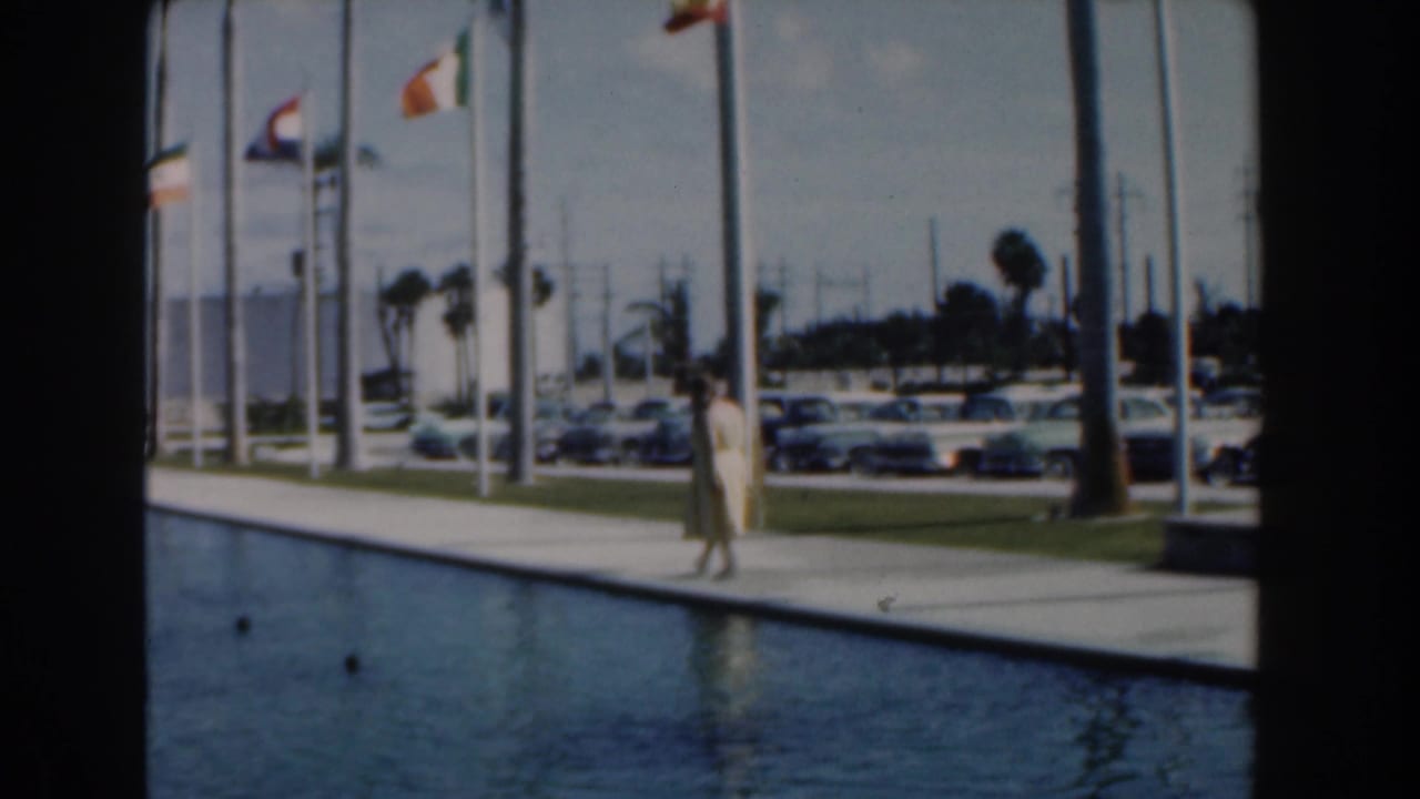 Woman walking near a long pool as flags, parked cars, poles, and palm trees line the background.