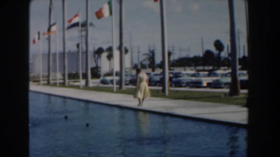 The woman appears closer to the center of the walkway beside the reflecting pool, with flags and mid-century cars behind her in Nassau.