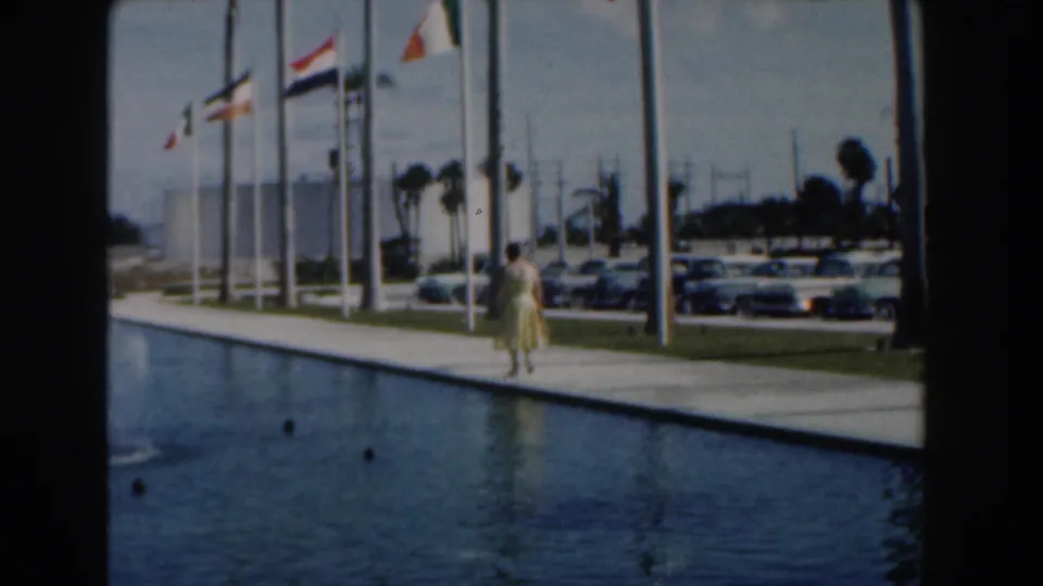 A final Nassau frame shows the woman farther down the poolside path, with flagpoles, parked classic cars, palms, and bright sky behind her.