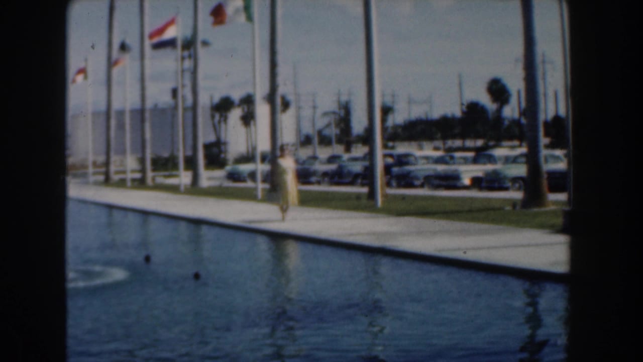 Woman in a yellow dress walks beside a long blue reflecting pool with flags and parked cars behind her.
