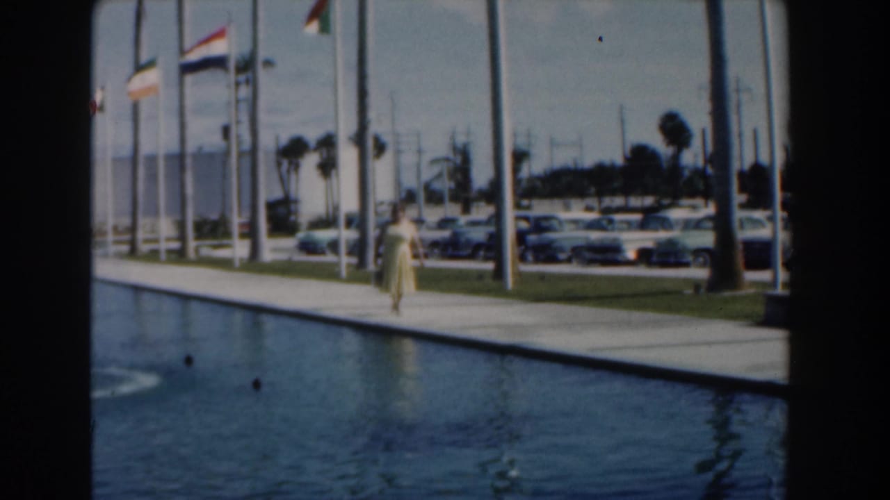 Woman continues along the poolside walkway as flags fly above parked cars and palm trees.