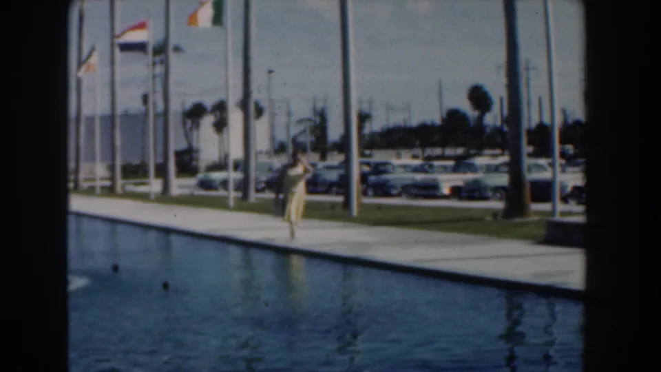 A 1961 Nassau travel frame shows the woman near the pool edge, with flags, cars, palms, and a building beyond.