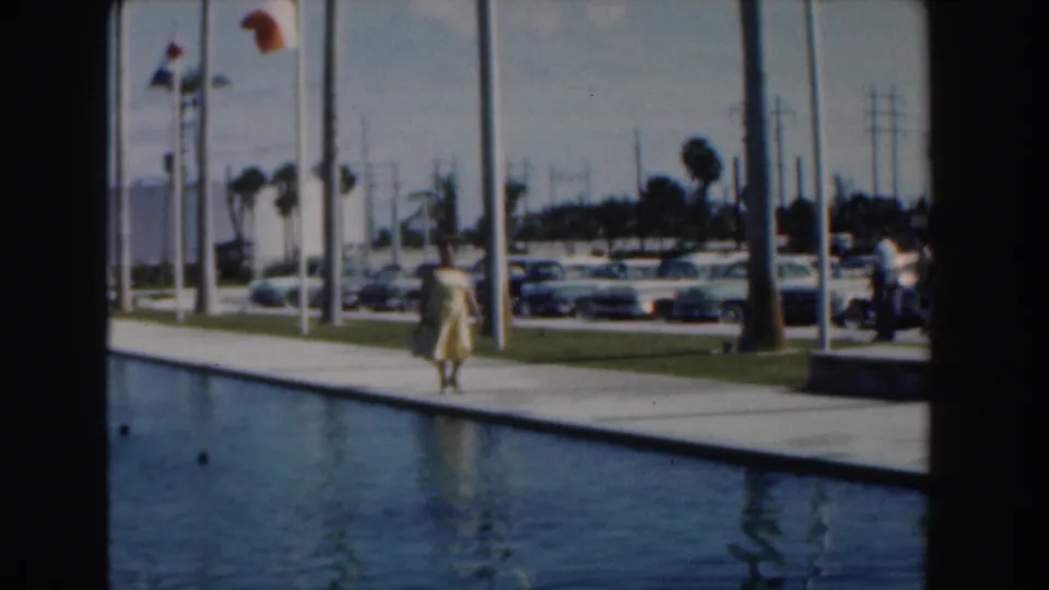 The final frame keeps the woman on the Nassau walkway beside the reflecting pool, with flags, cars, and palms behind her.