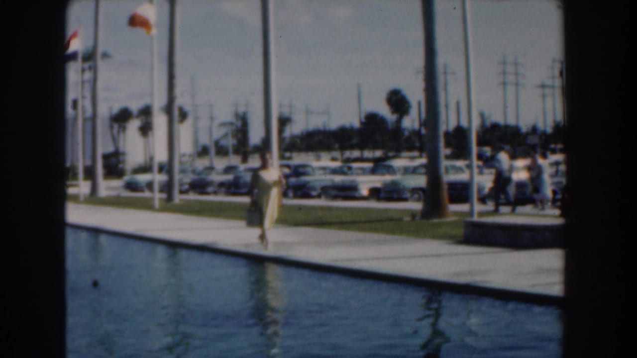Woman in a yellow dress walking beside a reflecting pool with cars and palm trees behind her.