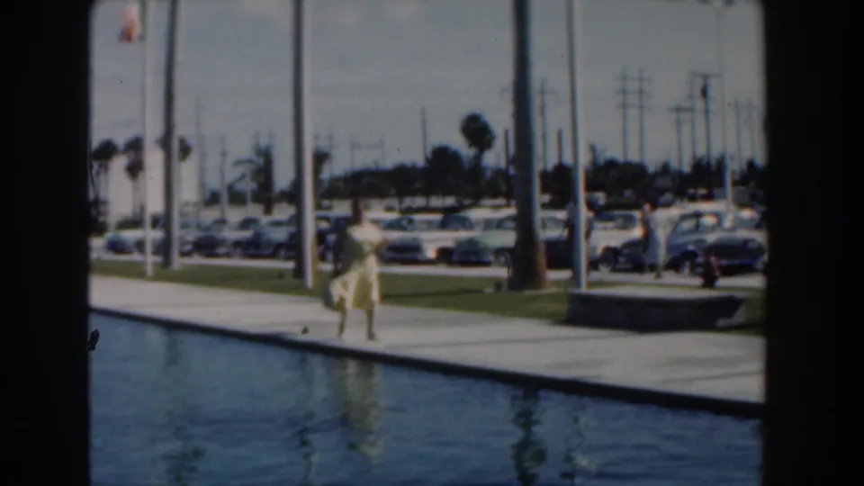 A woman in a yellow dress continues along the reflecting pool in Nassau, with flagpoles, palms, and a row of vintage cars beyond the walkway.