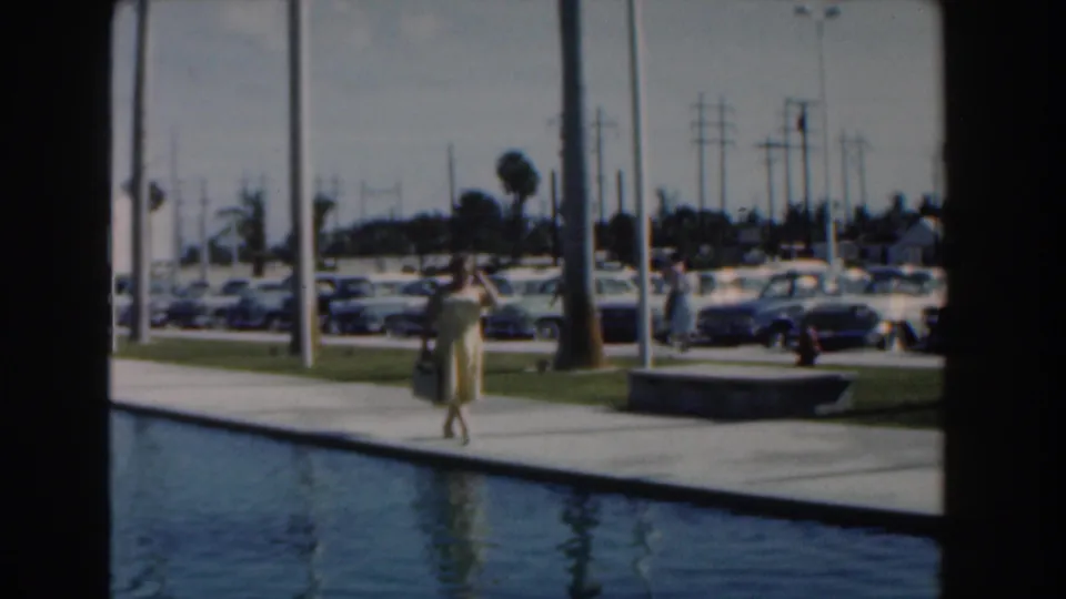The Nassau poolside scene shows the woman walking nearer the center of frame, with palms, flagpoles, vintage cars, and pedestrians in the distance.