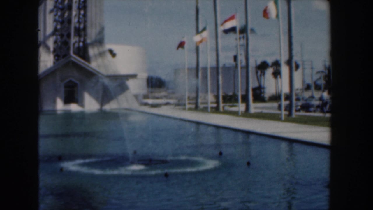 Reflecting pool with fountain spray, flagpoles, palm trees, and an angular building beyond.