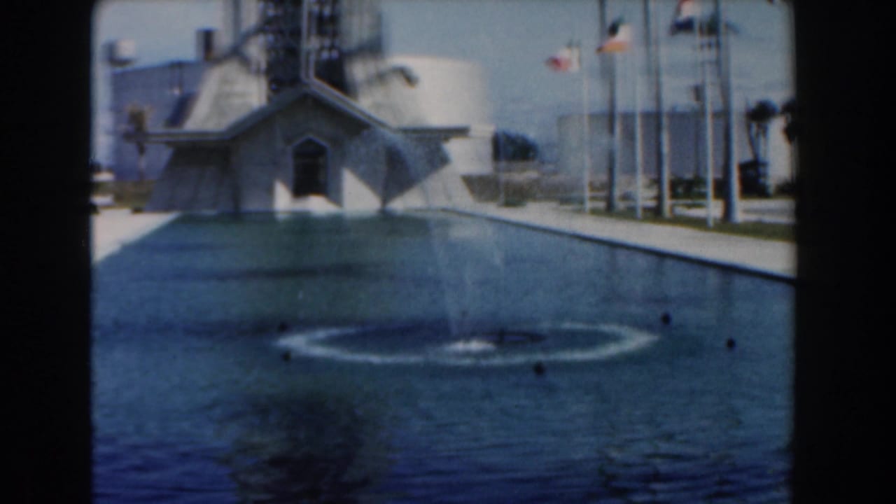 Fountain spray in a long pool facing an angular building with flags along the plaza.