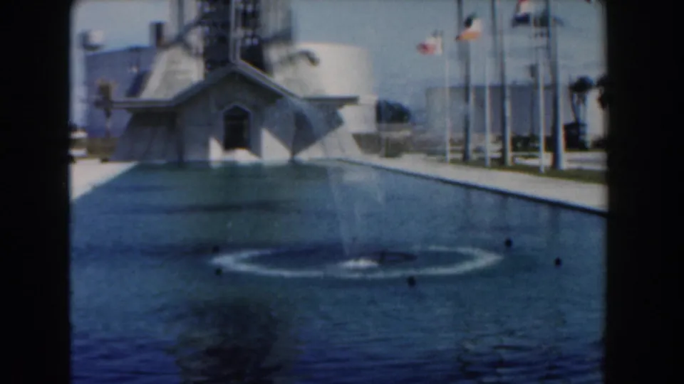 1961 Nassau frame looking across the reflecting pool toward an angular building, with flagpoles and palm trees around the plaza.
