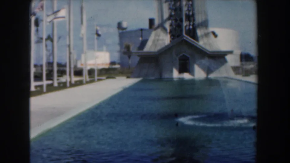 1961 Nassau home-movie view of a plaza walkway, flagpoles, reflecting pool, and angular landmark building under bright daylight.