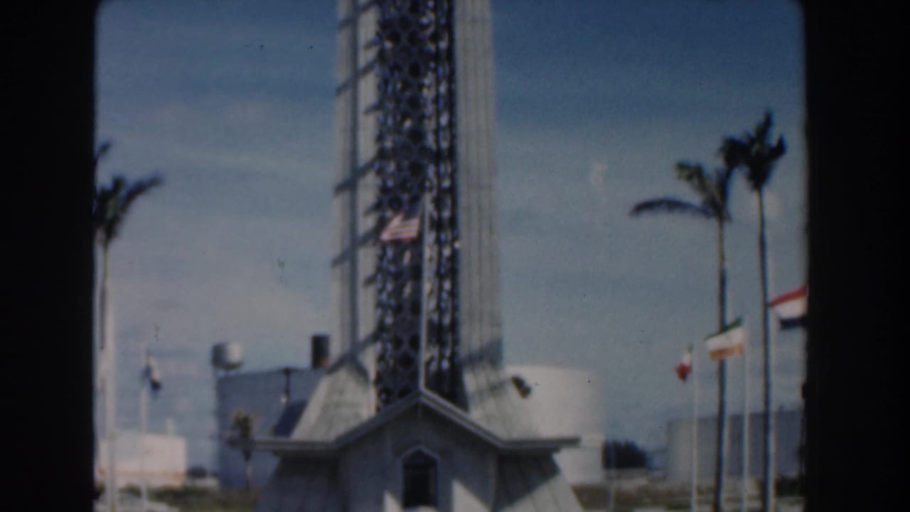 1961 Nassau home-movie frame showing the base of a tall modernist lattice tower with palms, flags, and bright sky.