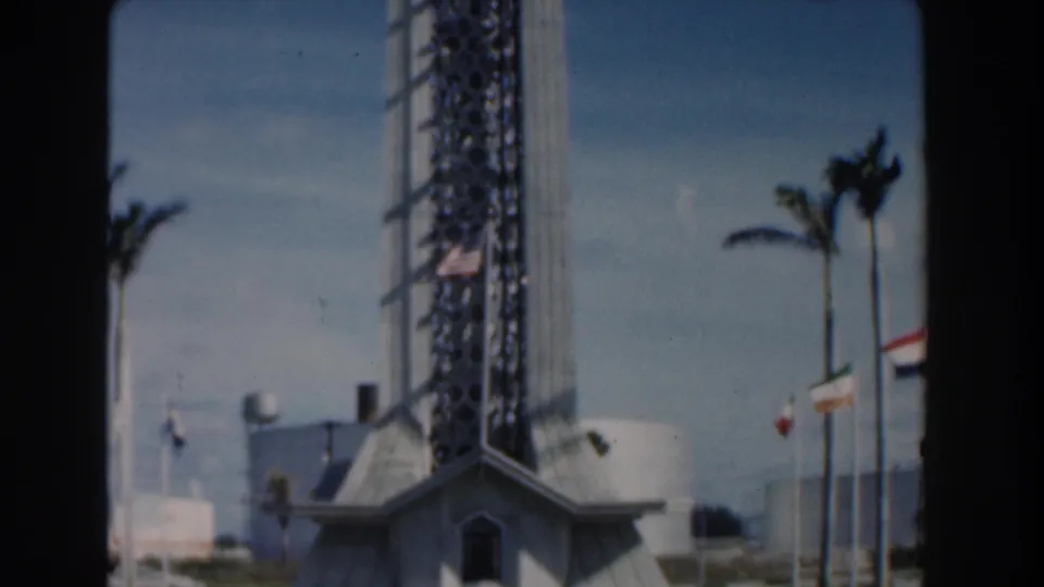 1961 Nassau home-movie frame showing the base of a tall modernist lattice tower with palms, flags, and bright sky.