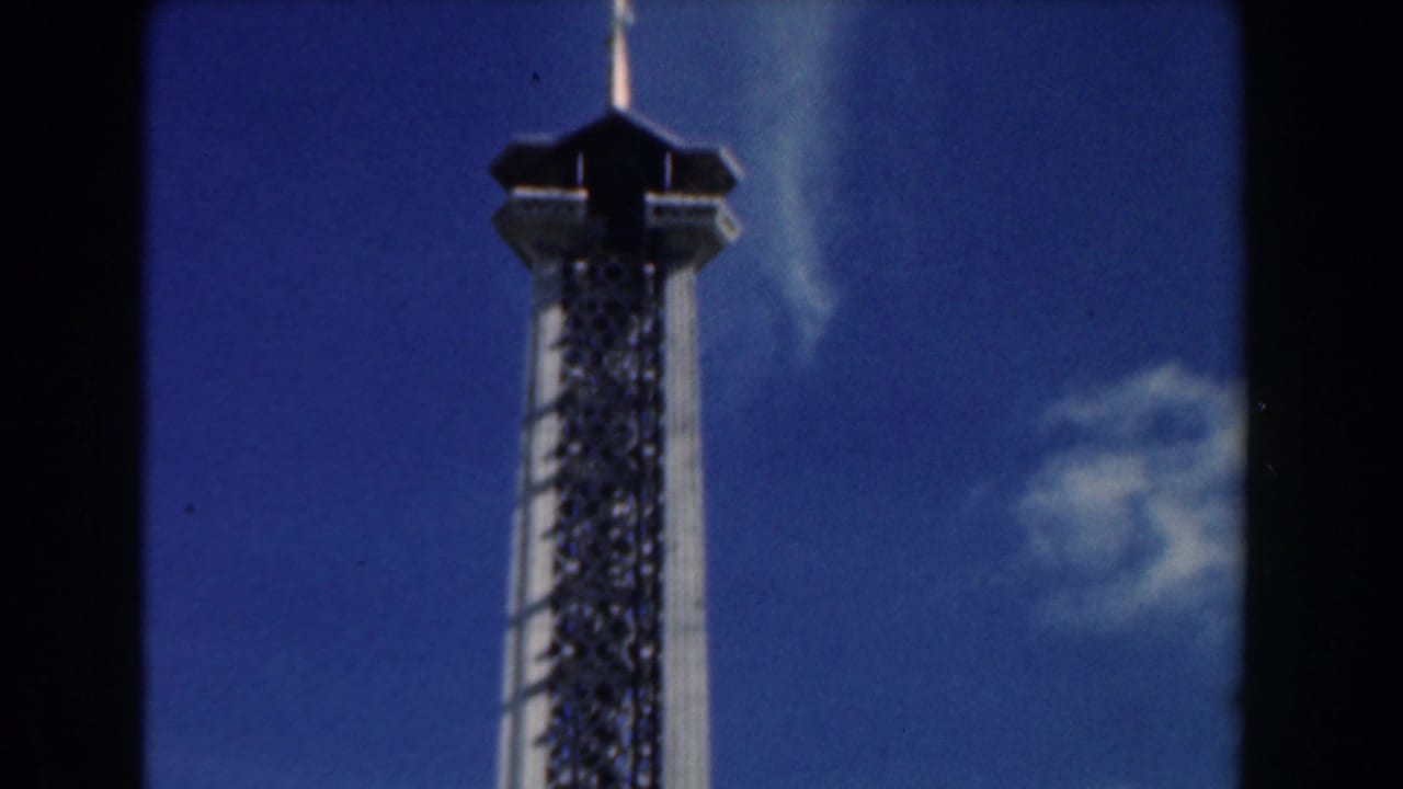 Low-angle 1961 Nassau 8mm view of a tall lattice attraction tower rising into a blue sky.