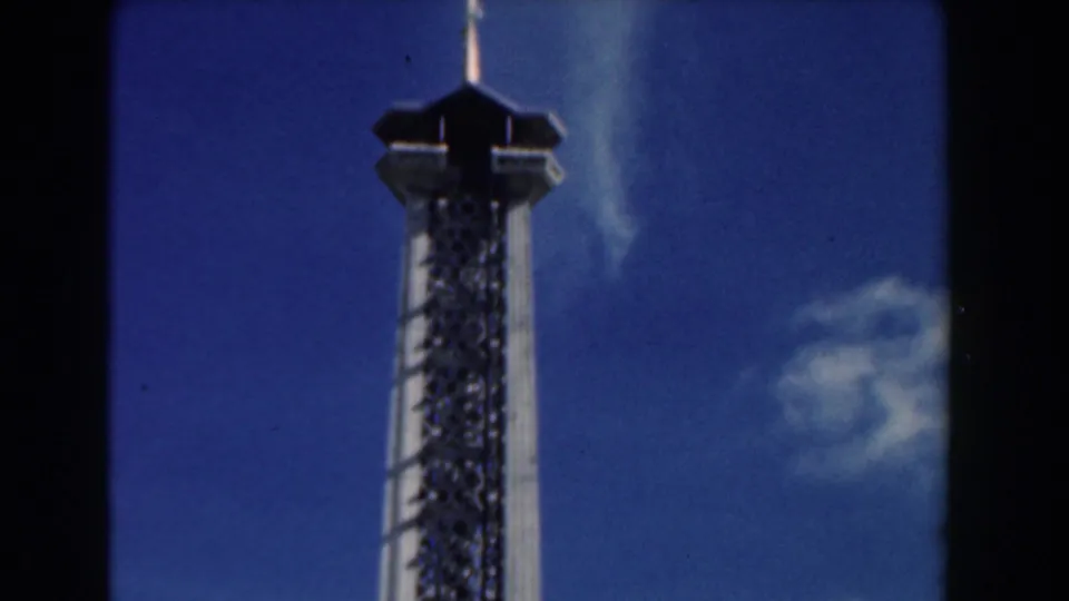 Low-angle 1961 Nassau 8mm view of a tall lattice attraction tower rising into a blue sky.