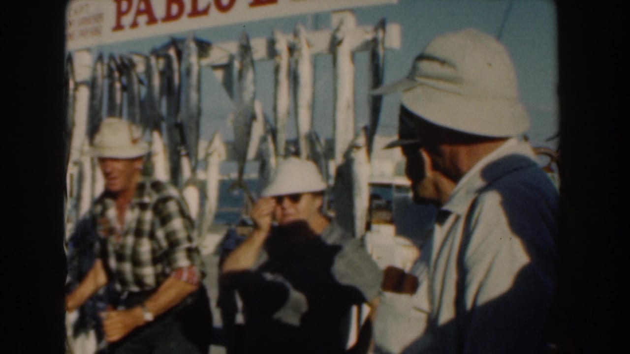 Adults in hats stand near a boat with fish hanging behind them.