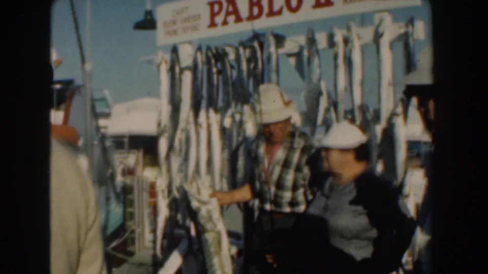 Nassau, Bahamas 1961 home-movie frame showing adults examining a hanging catch beside a boat at a fish-market scene.