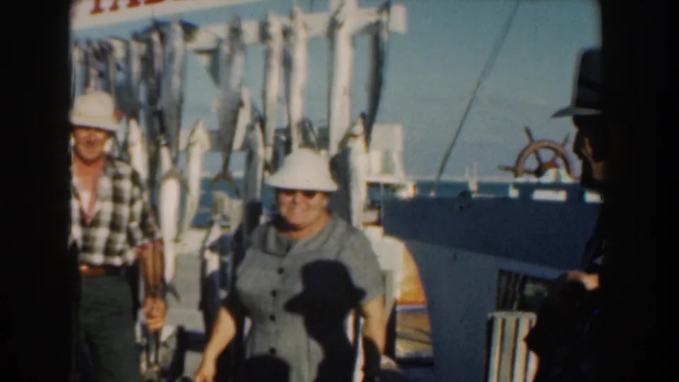 1961 8mm frame of adults on the Pablo II boat deck near hanging fish, with blue water and sky in the background.