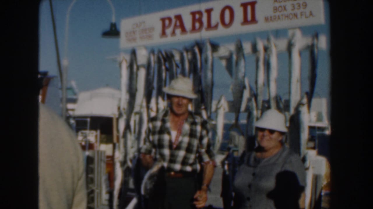 Two adults in white hats stand on boat stairs beneath a Pablo II sign and rows of hanging fish.