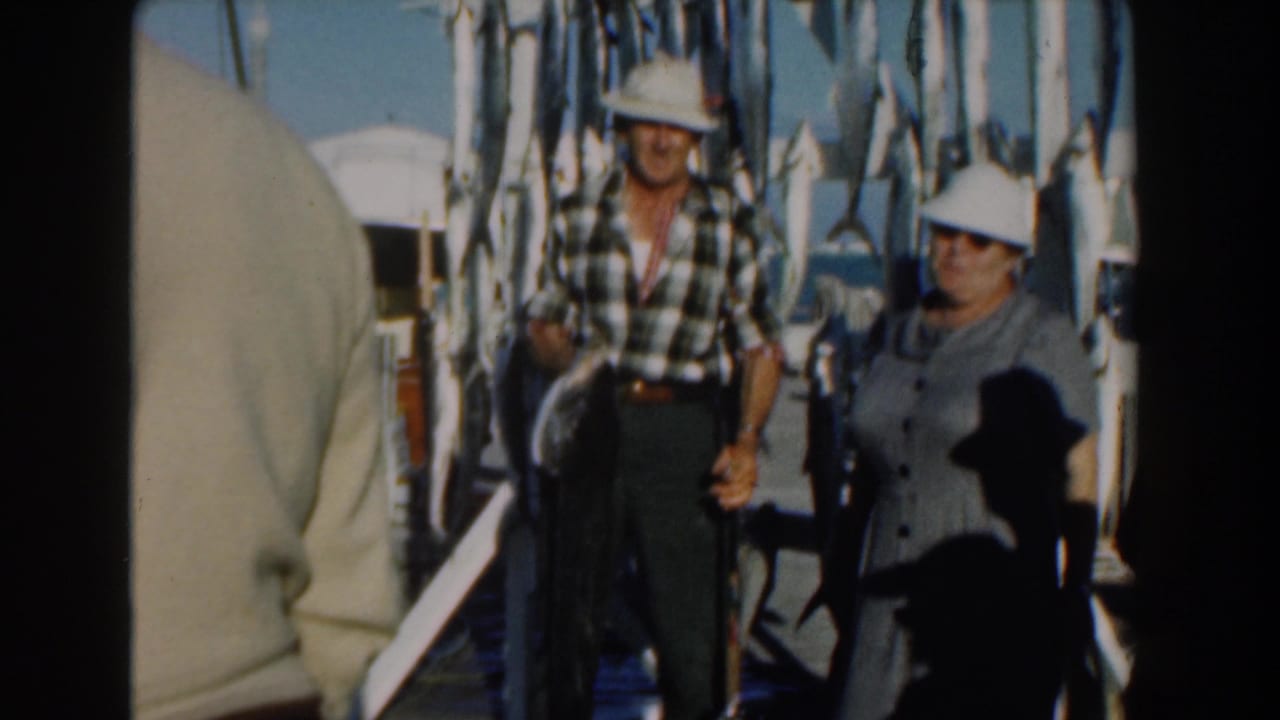 Man and woman in white hats stand near hanging fish, with a partial foreground figure at left.
