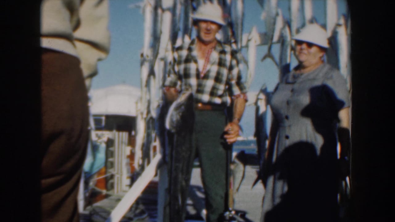 In 1961 Nassau, a man in a plaid shirt and white hat holds a large fish while a woman in a gray dress stands beside him on a dock.
