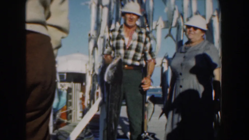 In 1961 Nassau, a man in a plaid shirt and white hat holds a large fish while a woman in a gray dress stands beside him on a dock.
