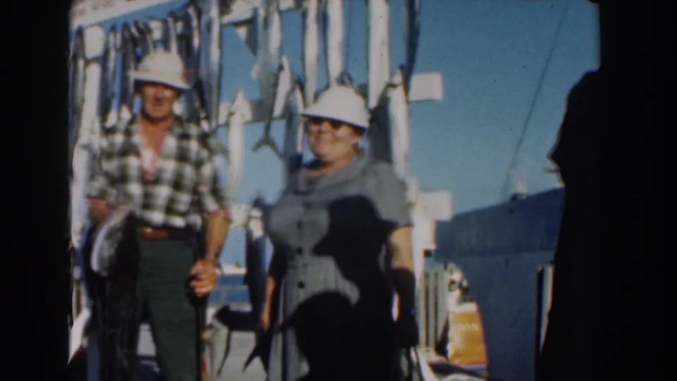 The Nassau dock scene continues with the man holding the catch upright and the woman standing close beside him under bright sun.
